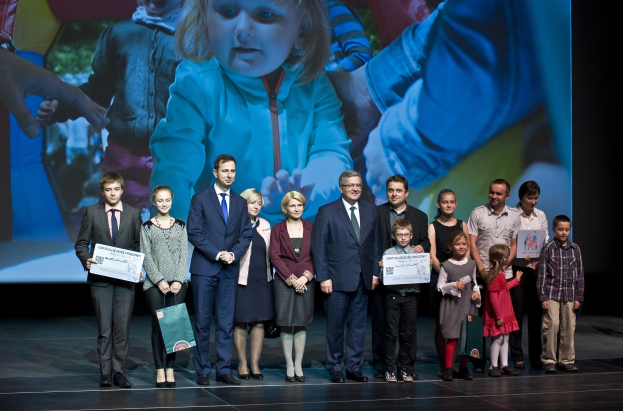 Eine Gruppe von Menschen auf einer Bühne, einige halten Taschen und Urkunden, mit einem Hintergrundbild auf einem Bildschirm.