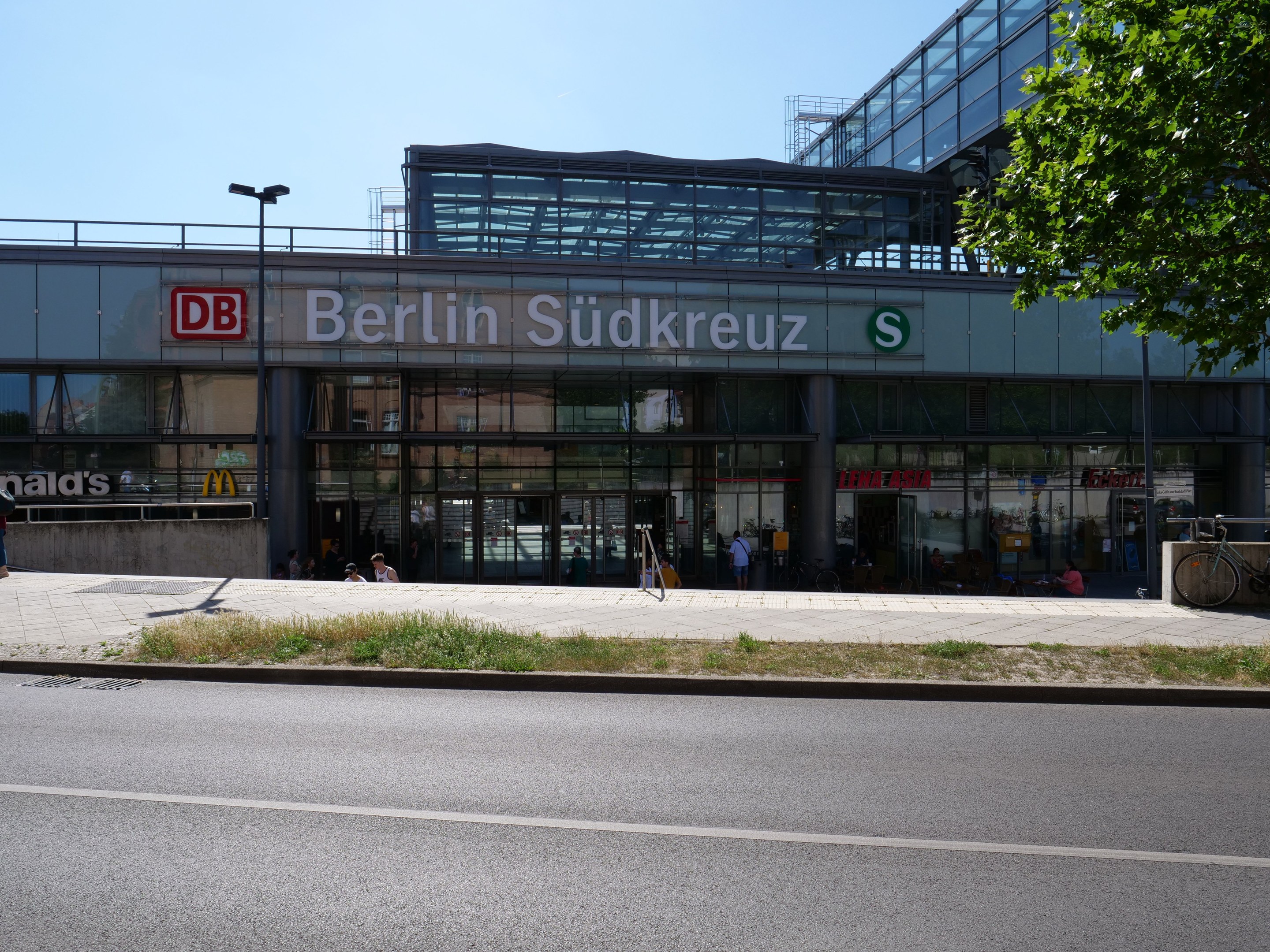 Eine große moderne Bahnstation mit Glaswänden, die Text anzeigen, umgeben von Straßeninfrastruktur, Fahrzeugen, Fußgängern, Fahrrädern, Bäumen und einem klaren Himmel.