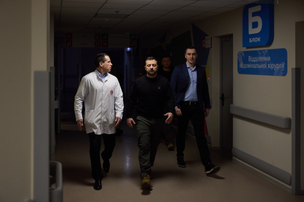 Eine Gruppe von Männern geht einen Krankenhausflur entlang, an dessen Decke Lampen hängen, vorbei an einer Tür auf der rechten Seite und Texttafeln auf der linken Seite, wobei sie besorgt wirken.