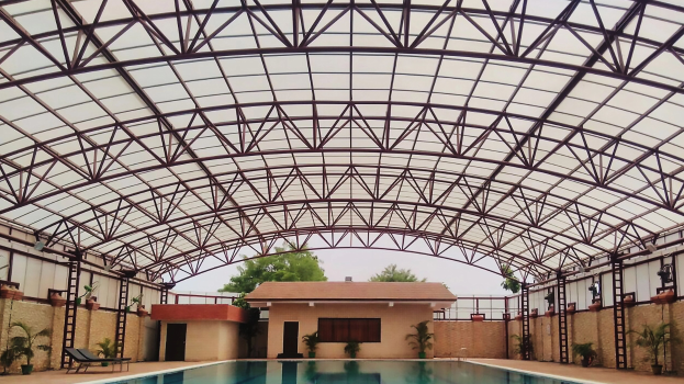 Großer Hallenbad mit Hauspflanzen, Wänden, Fenstern, Türen und Leitern umgeben, mit einem Dach aus Eisenstangen, Bäumen und einem klaren blauen Himmel im Hintergrund.