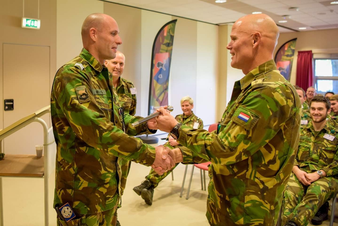 Zwei Männer in deutscher Militäruniform, die sich vor sitzenden Teilnehmern einer Innenveranstaltung mit Bannern, einem Tisch und Deckenleuchten die Hand geben.