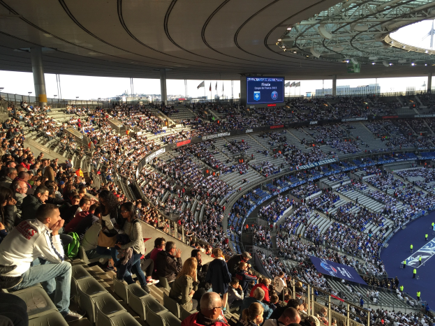 Großes Publikum in einem Stadion bei einem Fußballspiel, mit einer Bühne rechts, Fahnen, Stangen, einem Bildschirm und der Allianz Arena in München, Deutschland im Hintergrund.