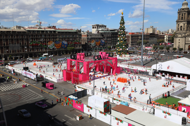Eine große Gruppe von Menschen auf einer Eisbahn in einer Stadt, umgeben von Gebäuden, Bäumen und Fahrzeugen, mit einem geschmückten Weihnachtsbaum im Hintergrund.
