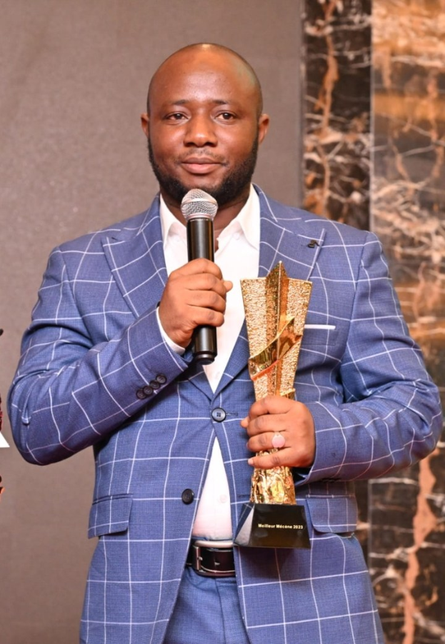 Ein Mann in einem blauen Anzug mit Mikrofon und Trophäe, der selbstbewusst und freudestrahlend steht, wahrscheinlich nach einem Award-Gewinn.