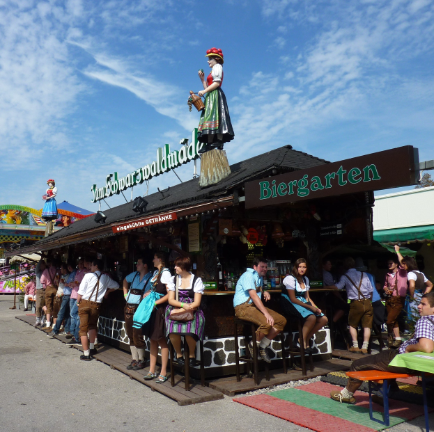 Gruppe von Menschen vor einem Gebäude auf dem Oktoberfest in München, einige in traditioneller bayrischer Tracht, andere auf Häckeln sitzend mit Tischen voller Flaschen und einer Tafel im Hintergrund, Bäume und bewölkter Himmel sichtbar.