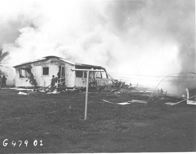Schwarzes und weißes Foto eines feuerbeschädigten Hauses mit Menschen davor, einem Vordergrundmast, Bäumen und Himmel im Hintergrund, mit Text unten.