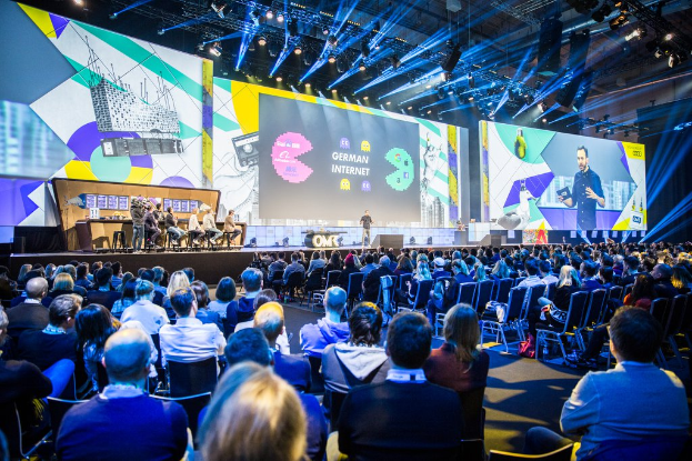 Eine große Menschenmenge auf Stühlen sitzend, die einer Bühne zugewandt ist, mit einem Tisch voller Gegenstände auf der linken Seite und Bildschirmen und Lichtern im Hintergrund.