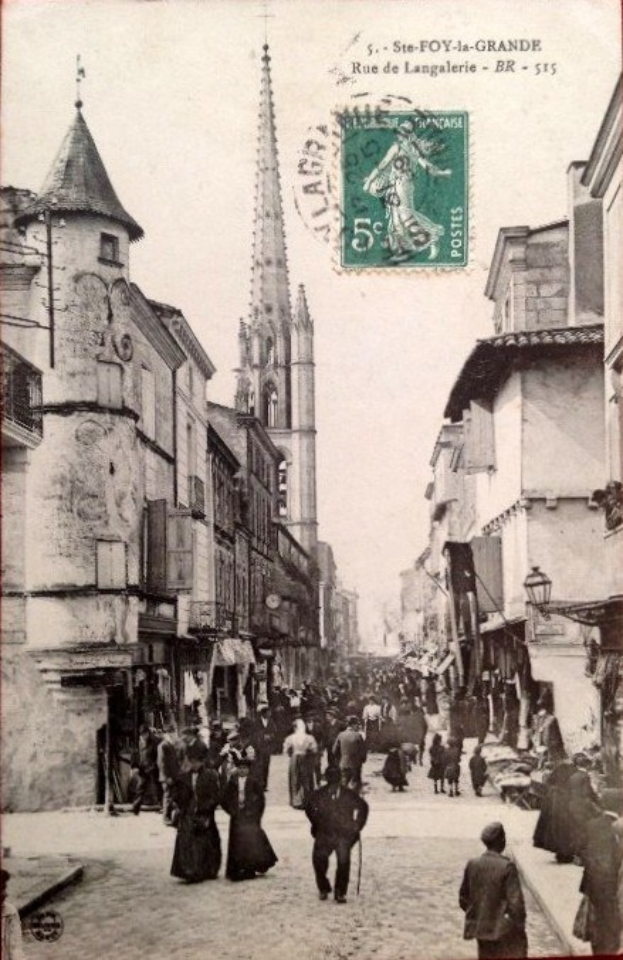 Altes Schwarz-Weiß-Foto von Fußgängern auf einer Stadtstraße mit Gebäuden auf beiden Seiten und einer zentralen Figur in der Menge sowie einem Stempel und Text oben.