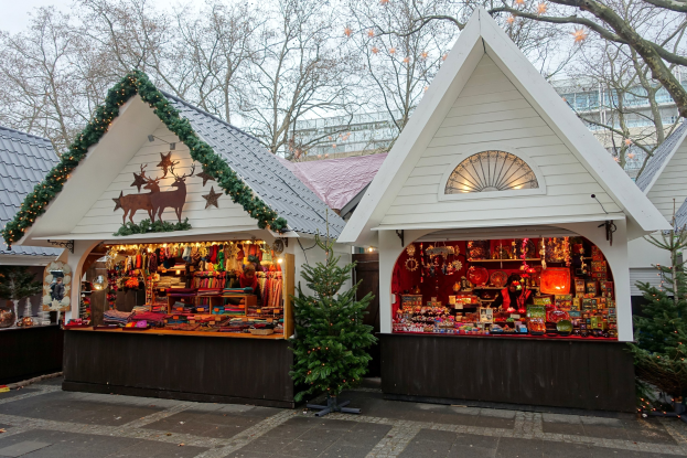 Ein kleines weißes Gebäude mit einem Weihnachtsmarkt davor, umgeben von Häusern, Bäumen und einem klaren blauen Himmel, mit festlichen Dekorationen und Lichtern im Inneren.