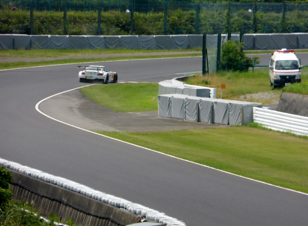 Ein Rennwagen fährt auf einer Rennstrecke mit einem Polizeiwagen im Hintergrund, umgeben von Gras, Pflanzen, Bäumen und Zaun, mit einem Fahrzeug auf der rechten Seite und Text unten.