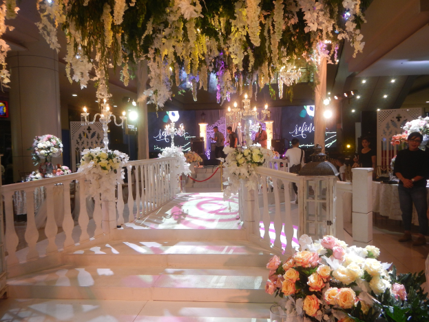 Hochzeitszeremonie in einem großen, prunkvoll dekorierten Raum mit weißen Blumen, Kronleuchtern, Geländern und Blumenschmuck, bei der Menschen auf dem Boden stehen und an Tischen sitzen.