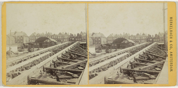 Schwarz-weiß-Foto einer Bahnschiene mit Gebäuden im Hintergrund, Text 'Merkelbach & Co, Co. Amsterdam' rechts und hölzernen Gegenständen im Vordergrund verstreut.