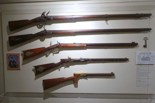 Vitrine mit verschiedenen Feuerwaffen, darunter ein Steinschlossgewehr, mit einem gerahmten Foto links und Dokumenten rechts.