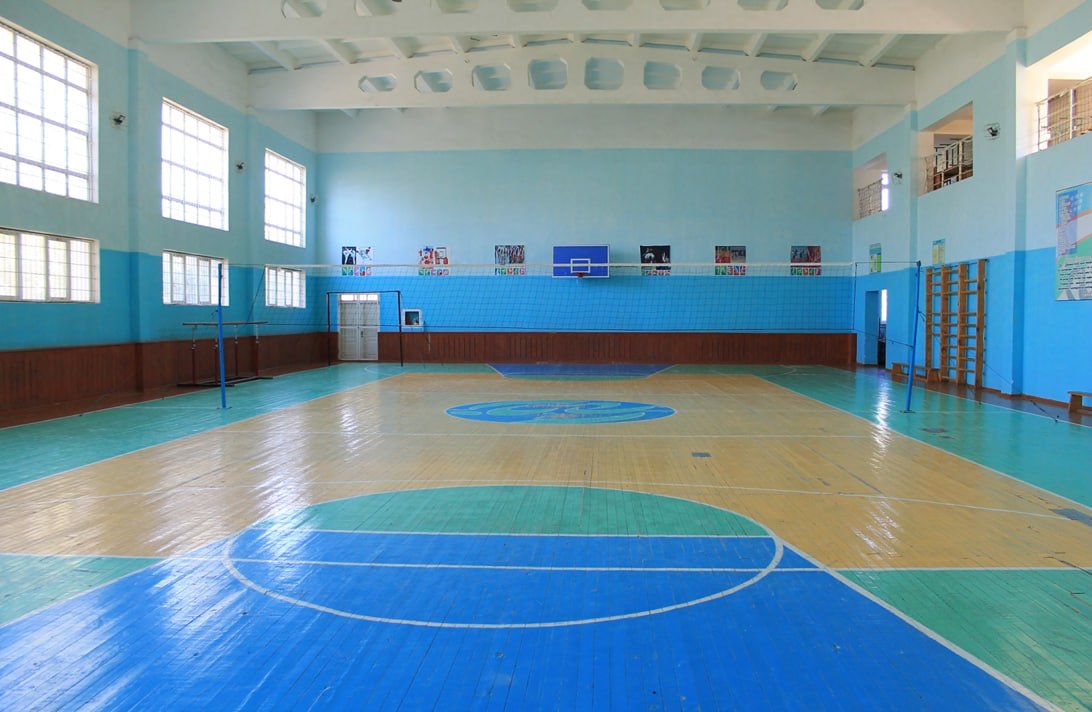 Großer Turnhalle mit einem blauen und weißen Basketballfeld, Netz, Pfosten, einer Tür, Fenstern, Plakaten an den Wänden, einem Geländer und einem Dach.