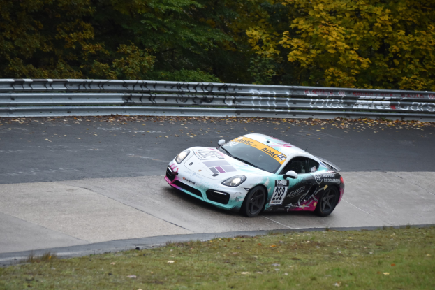 Ein Porsche Cayman GT4 fährt auf der Nürburgring 24 Stunden-Strecke, umgeben von grünem Gras und Bäumen, mit einem Geländer an der Seite.