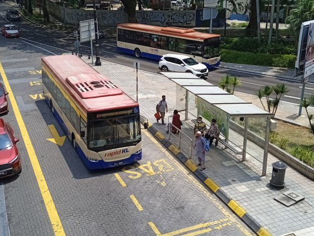 Stadtstraße mit fahrenden Autos und Bussen, eine Bushaltestelle auf dem Gehweg, Fußgänger auf dem Gehweg und umliegende städtische Elemente einschließlich Pfosten, Beschilderung, Bäume, Gras und Gebäude.