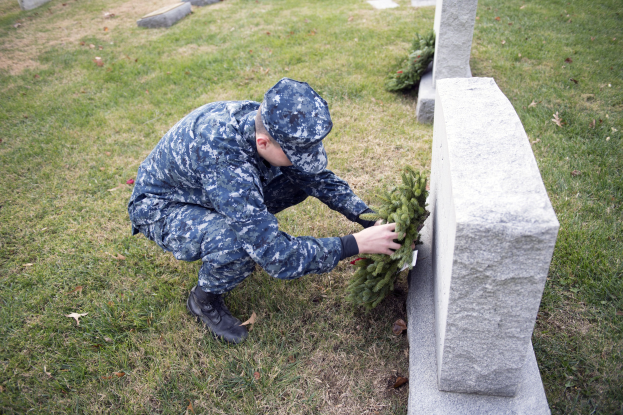 Ein Mann in militärischer Uniform kniet, um einen Kranz auf ein Grab zu legen, mit anderen Gräbern im Hintergrund.