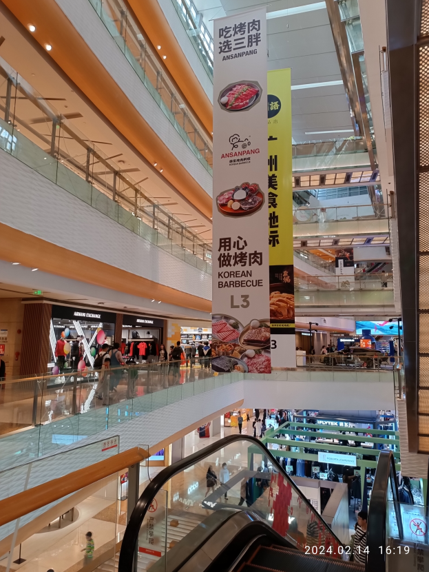 Ein Innenblick in ein Einkaufszentrum mit einer Rolltreppe in der Mitte, Menschen, die herumlaufen, einige mit Handtaschen, und Schilder, die von der Decke hängen, mit Geschäften im Hintergrund.