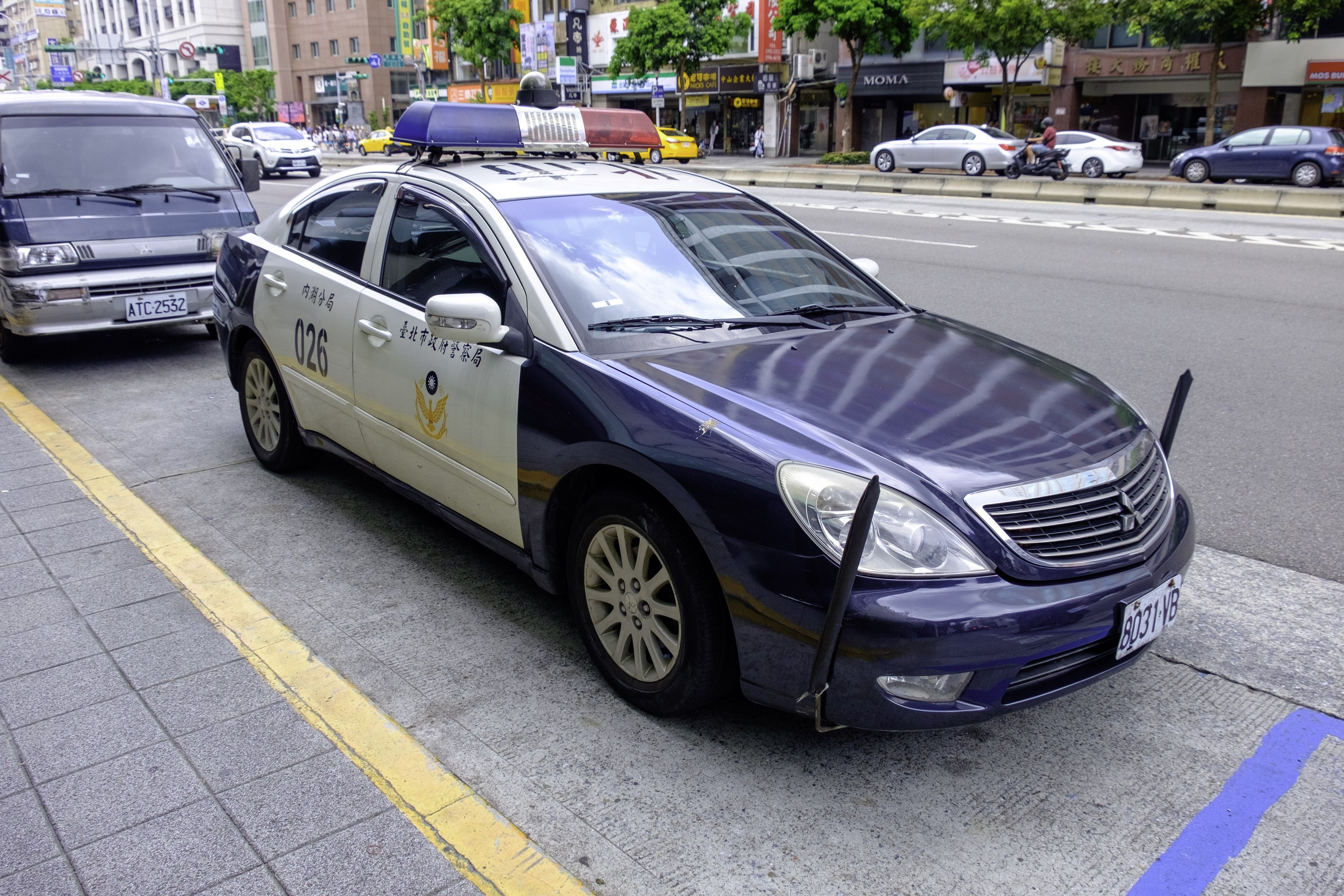 Ein Polizeiwagen am Straßenrand mit Gebäuden, Bäumen, Strommasten, Schildern, Werbetafeln und Menschen in der Nähe, dessen Vorderrad sichtbar ist.
