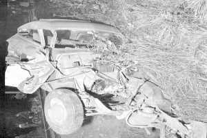 Schwarzes und weißes Foto eines schwer beschädigten Autos mit eingedellter Front und zersplitterten Fenstern am Straßenrand, mit einem Baum und verstreuten Gegenständen im Hintergrund.