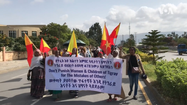Eine Gruppe von Menschen marschiert auf einer Straße, hält Fahnen und ein Banner mit Text, mit Pflanzen und einem Fahrzeug auf der rechten Seite und einem Gebäude, Bäumen, Pfählen, einem Turm und dem Himmel im Hintergrund.