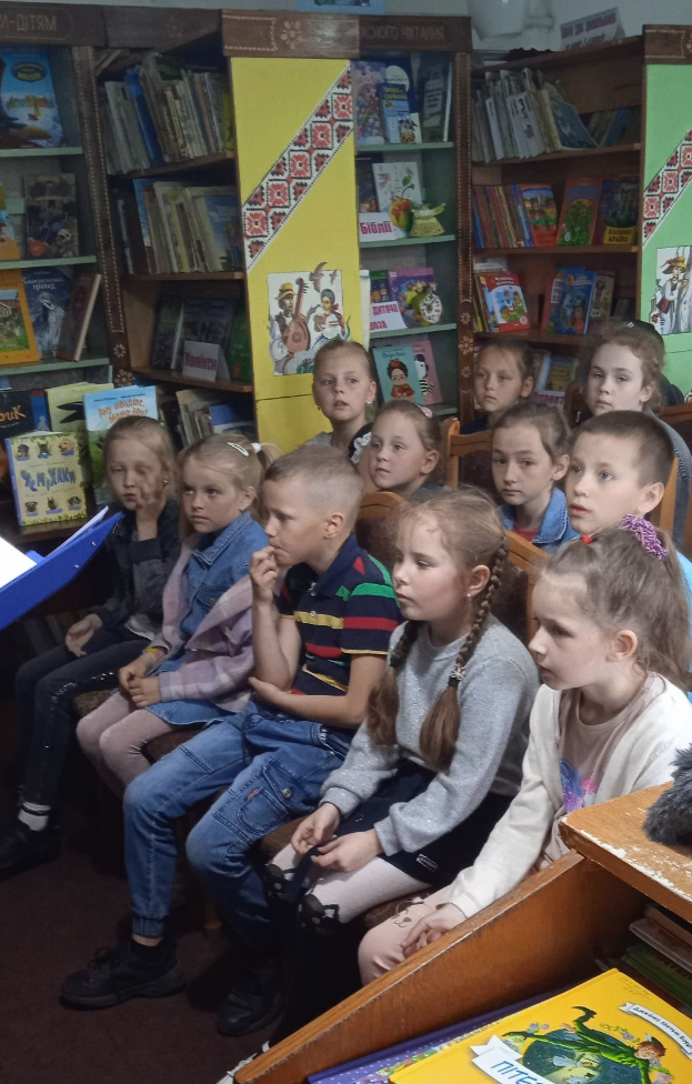 Eine Gruppe von Kindern sitzt in einer Bibliothek umgeben von Bücherregalen, mit einigen Lesen auf Tischen auf beiden Seiten.