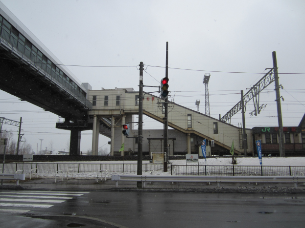 Eine Bahnhofs-Szene mit einem Zug auf Schienen, einer Brücke mit Pfeilern und Geländern, Strommasten mit Drähten, Verkehrssignalen, Geländern mit Tafeln, schneebedeckter Erde, Bäumen und einem klaren Himmel.