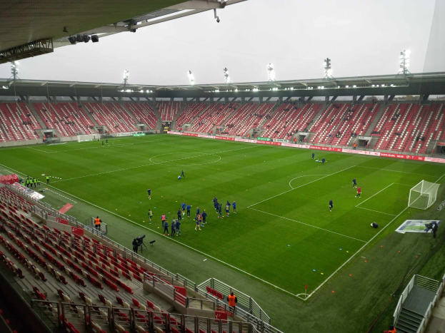 Ein Fußballfeld in einem Stadion mit einer Gruppe von Menschen, umgeben von Sitzplätzen, Geländern, Stufen und Lichtern, mit dem Himmel im Hintergrund.