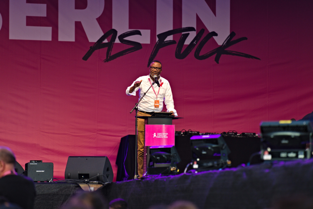 Ein Mann in einem weißen Hemd und braunen Hosen steht auf einer Bühne und hält ein Mikrofon in der Hand, während er eine Menge anspricht. Im Hintergrund hängt ein pinkfarbenes Banner mit der Aufschrift "Berlin as Fuck".