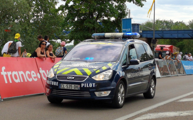 Polizeifahrzeug fährt an einer Menge vorbei, die Schilder, Bäume, eine Brücke, eine Flagge und einen bewölkten Himmel im Hintergrund hat.