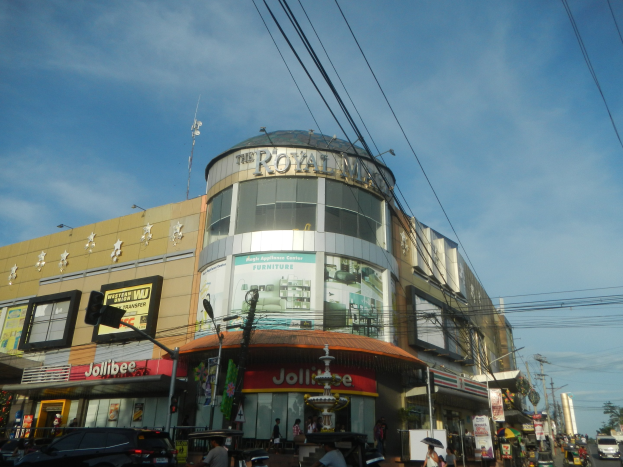 Eine belebte Stadtstraße mit fahrenden Fahrzeugen, Fußgängern auf dem Gehweg und dem Royal Mall Einkaufszentrum im Hintergrund, umgeben von Bäumen, Strommasten und Drähten unter einem Himmel.