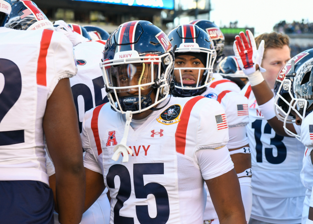 Eine Gruppe von Männern in Helmen und Handschuhen steht zusammen auf einem Feld, mit einer unscharfen Menge, einem Schild mit der Aufschrift "Auburn vs Virginia Tech - 11/28/15 College Football Pick, Odds, and Prediction" und einem Himmel im Hintergrund.