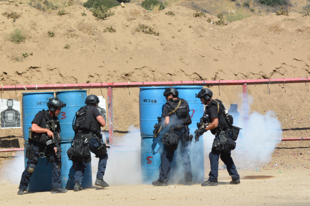 Eine Gruppe von Polizisten in schwarzer Einsatzausrüstung mit Waffen auf einem Feld mit blauen Tonnen, einem roten Zaun, einem Plakat, Pflanzen und einem Berg im Hintergrund.