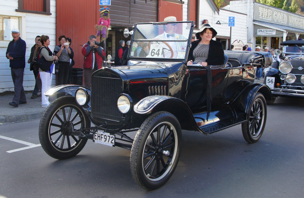 Eine Frau fährt ein Oldtimer-Auto eine Straße mit Gebäuden, Bäumen und Schildern entlang, während Menschen mit Kameras auf dem Gehweg unter einem sichtbaren Himmel stehen.