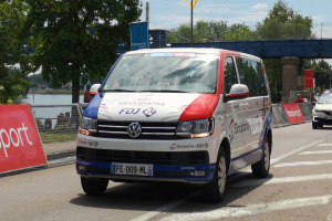 Ein weißer und roter Lieferwagen mit einem sichtbaren Rad fährt auf einer Straße neben einer Brücke, mit einem Geländer auf der linken Seite, Textbannern, Bäumen im Hintergrund und einem bewölkten Himmel.