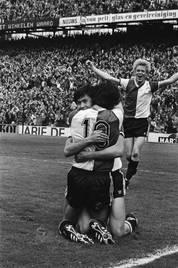 Drei Fussballspieler umarmen sich auf dem Boden nach einem Torerfolg, mit jubelnder Menge und schwenkenden Fahnen im Hintergrund, in einem schwarz-weiss gestalteten Stadion.