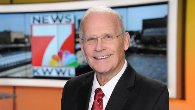 Mann im Anzug und Krawatte mit Brille vor einem Fernseher stehend und l├Ąchelnd, wahrscheinlich ein Nachrichtensprecher.