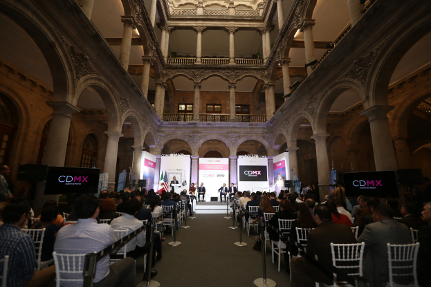 Eine große Gruppe von Menschen, die in Stühlen in einem Gebäude sitzen, mit einer zentralen Bühne, die von Ständen mit Polen und Bannern, Säulen, Geländern, Fenstern und Deckenleuchten flankiert wird, was auf ein Ereignis in New York City hindeutet.