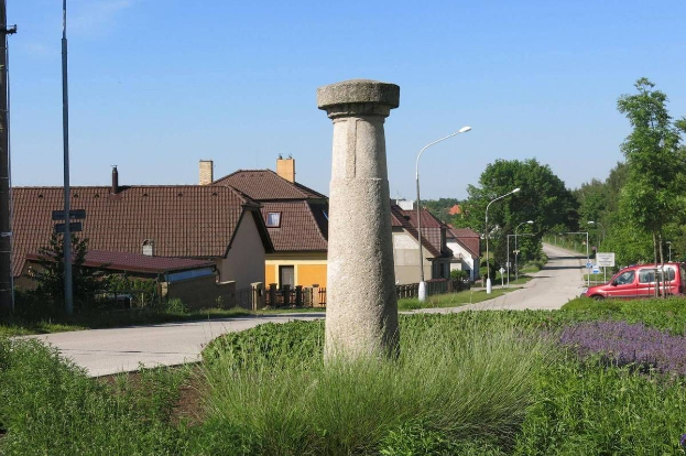 Steinsäule in einer grünen Wiese mit umliegenden Pflanzen und einem Straßenabschnitt mit Fahrzeugen, vor Häusern und Himmel.