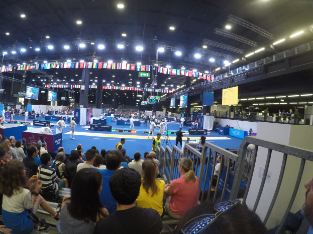 Große Menschenmenge sitzend und stehend in einer Indoor-Sportarena mit Bildschirmen, Fahnen und Deckenbeleuchtung im Hintergrund.