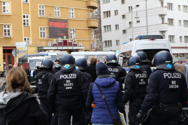 Polizeibeamte in Uniform vor einer Menge während einer Demonstration in Berlin, mit Fahrzeugen, Gebäuden und einer Person mit einer Kamera im Bild.