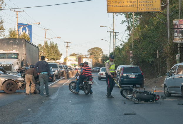 Gruppe von Menschen um ein verunglücktes Motorrad auf der Straße herumstehend mit mehreren Fahrzeugen, darunter ein Lastwagen, und Hintergrund-Elementen wie Bäume, Pfosten, Lampen, Schilder und Himmel.