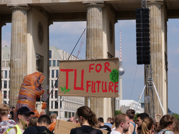 Eine Gruppe von Menschen vor dem Brandenburger Tor in Berlin, Deutschland, mit einigen, die Taschen tragen und einer in Kostüm mit einem Schild, vor einem Hintergrund von Gebäuden, Bäumen und einem Turm unter einem bewölkten Himmel.