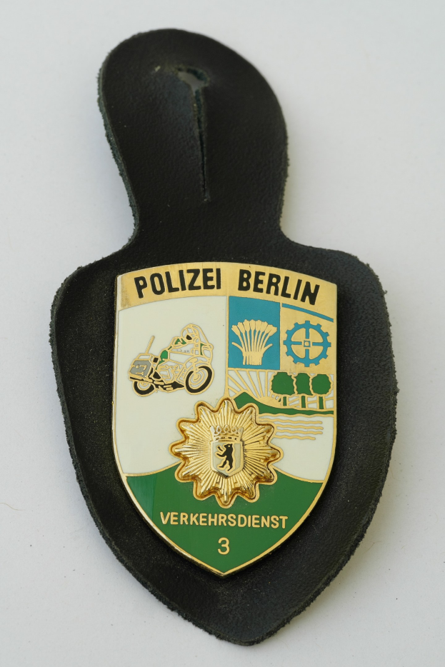 Ein schildförmiges Polizeibadge mit der Aufschrift "Polizei Berlin" auf einem blauen Hintergrund, einem weißen Rand, einem weißen zentralen Stern und schwarzer fetter Schrift, das auf einer Oberfläche liegt.