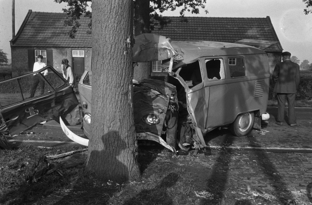 Schwarz-weißes Bild eines verunglückten Autos am Straßenrand mit einem Baum im Vordergrund, Menschen in der Nähe und Häusern, Bäumen und einem Pfahl im Hintergrund.