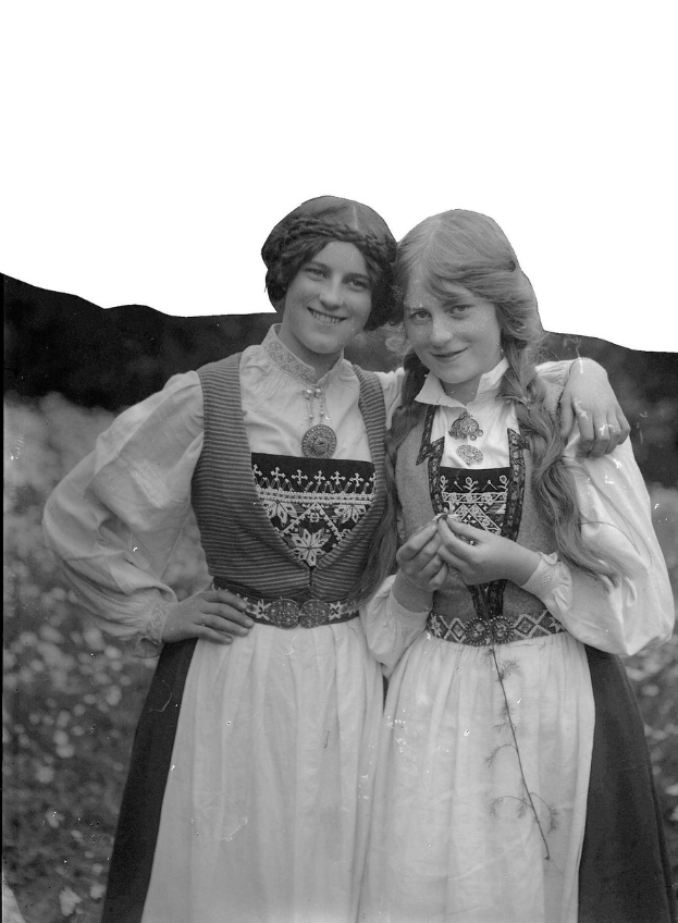 Zwei junge Frauen in traditioneller bayrischer Tracht stehen lachend, tragen weiße und schwarze bestickte Kleider mit Perlen, vor einem Hintergrund aus grünen Pflanzen und einem hellen Himmel.