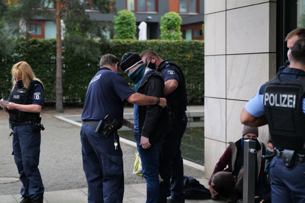 Eine Gruppe von Polizisten steht vor einem Gebäude, während eine Frau in der Nähe ein Buch und einen Stift hält und andere im Hintergrund auf dem Boden sitzen oder stehen, umgeben von Pflanzen, Bäumen und Gebäuden.