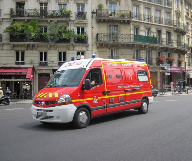 Rettungswagen auf der Straße vor einem Gebäude mit Fenstern, Geländern, Pflanzen und anderen Gegenständen geparkt, mit ein paar Menschen und Fahrrädern im Hintergrund.