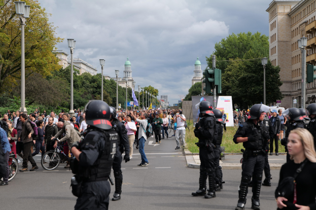Eine Gruppe von Menschen geht eine Straße entlang mit Polizisten in Helmen und bewaffnet, neben Fahrrädern, Laternen, Verkehrsampeln, Bäumen und Gebäuden unter einem bewölkten Himmel.