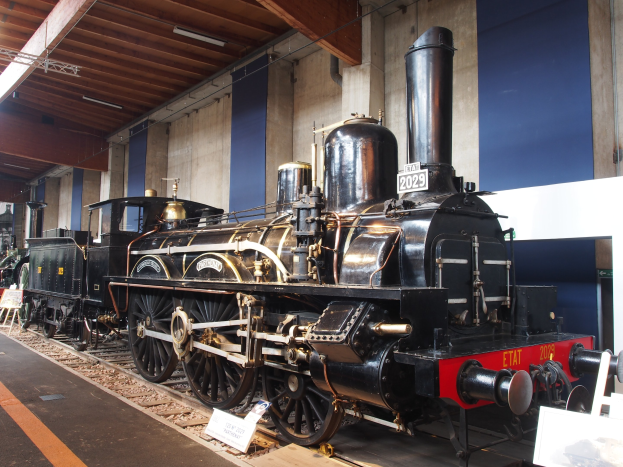 Eine alte Dampflokomotive in einem Museums Setting umgeben von Eisenbahnschienen, Informationsschildern und anderen Ausstellungen, mit einer beleuchteten Decke und Wand im Hintergrund.